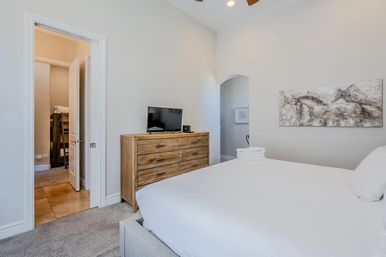Bright modern bedroom with crisp white bedding, rustic wooden dresser topped by a flat-screen TV, abstract horse wall art, arched doorway and neatly folded towels.