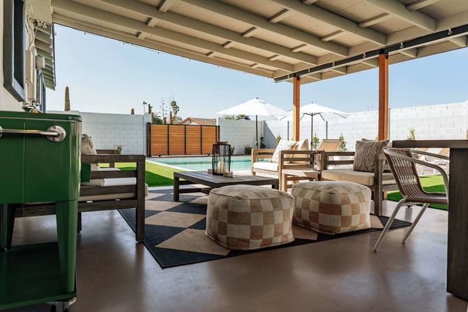 Covered backyard patio with wooden columns and modern outdoor lounge seating, checkered poufs and coffee table overlooking a private swimming pool with white privacy wall, sun umbrellas and clear blue sky.