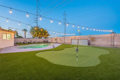 Dusk residential backyard with a synthetic putting green and checkered flags, curved swimming pool and lounge chairs, warm string lights overhead and palm trees against a blue sky.