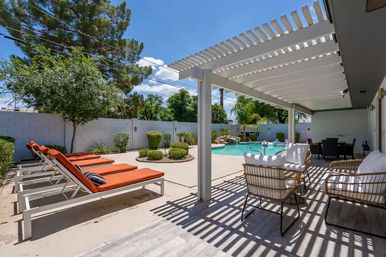 Sunny backyard pool patio with white pergola, wicker seating and orange-cushioned lounge chairs beside a curved swimming pool and landscaped yard.