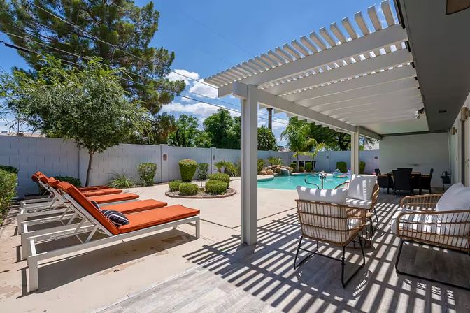 Sunny backyard pool patio with white pergola, wicker seating and orange-cushioned lounge chairs beside a curved swimming pool and landscaped yard.