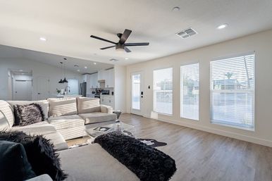 Sunlit modern open-concept living room and kitchen with beige sectional sofa, black accent pillows, glass coffee table, black ceiling fan, and light wood-look floors