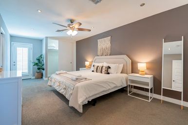 Cozy modern master bedroom with king bed and neutral linens, macrame wall hanging above an upholstered headboard, matching nightstands with lamps, ceiling fan, full-length mirror, potted plant, and French door with shutters.