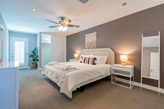 Cozy modern master bedroom with king bed and neutral linens, macrame wall hanging above an upholstered headboard, matching nightstands with lamps, ceiling fan, full-length mirror, potted plant, and French door with shutters.
