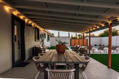 Covered backyard patio with a long rustic wood dining table and wicker chairs under warm string lights, overlooking a poolside lounge area and green turf at dusk