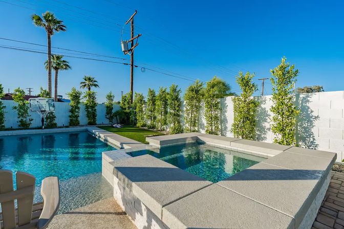 Sunny Southern California backyard with turquoise pool and raised spa, white block wall lined with tall hedges, palm trees, a basketball hoop and utility pole under a clear blue sky