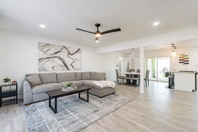 Modern open-concept living room and kitchen with a gray sectional sofa, abstract wall art, black ceiling fan, patterned area rug, wood‑look tile flooring, dining table, and kitchen island with bar stools by sliding patio doors.