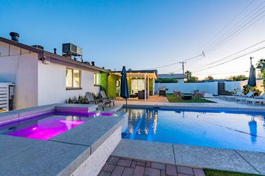 Evening suburban backyard with illuminated purple hot tub spilling into a blue swimming pool, lounge chairs, covered patio and outdoor seating.
