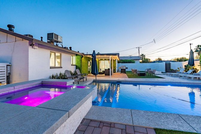 Evening suburban backyard with illuminated purple hot tub spilling into a blue swimming pool, lounge chairs, covered patio and outdoor seating.