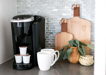 Black single-serve coffee maker with stacked pods and two white mugs on a modern kitchen countertop, backed by mosaic tile, wooden cutting boards and a small eucalyptus arrangement, ready for morning brew.