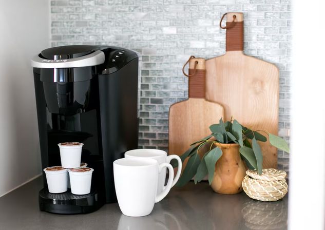 Black single-serve coffee maker with stacked pods and two white mugs on a modern kitchen countertop, backed by mosaic tile, wooden cutting boards and a small eucalyptus arrangement, ready for morning brew.