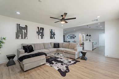 Bright modern open-plan living room and kitchen in a contemporary home — light-gray L-shaped sectional with black accent pillows, clear acrylic coffee table on a black-and-white cowhide rug, abstract monochrome wall art, dark ceiling fan, pendant lights and light wood floors.