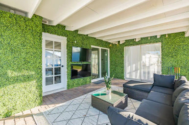 Sunny backyard patio with white beamed ceiling, artificial green hedge wall with two wall-mounted TVs, sliding glass doors, dark wicker sectional and glass-top coffee table on a patterned rug over paver flooring — cozy outdoor living space.