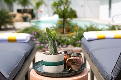 Poolside scene with a tiny spiky cactus in a striped ceramic pot on a tray with hammered copper mugs set between two cushioned lounge chairs with striped towels by a sunlit pool and flowering landscaping