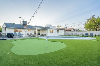 Suburban backyard at dusk with a synthetic putting green and flags, kidney-shaped swimming pool with tiled edge, patio lounge seating and string bistro lights overhead.