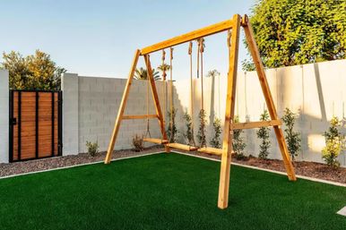 Wooden A-frame swing set with rope and bench seats on vibrant artificial turf in a sunny backyard with a white cinderblock privacy wall, young shrubs, wooden gate and palm trees in the background.
