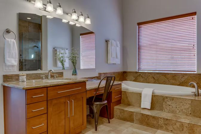 Bright master bathroom with wooden vanity and granite countertop, makeup nook with metal chair, large mirror and lights, tiled soaking tub with steps and wood blinds