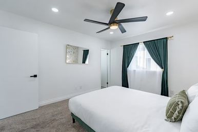 Contemporary bedroom interior: bright white walls, gray carpet, low-profile bed with white linens and green throw pillow, black five-blade ceiling fan, deep green curtains framing a sunlit window.