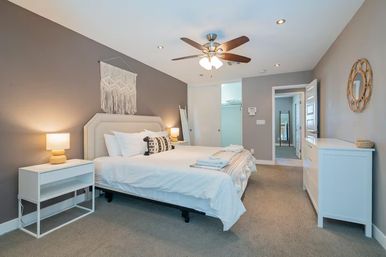 Bright modern master bedroom with king bed and white linens, macrame wall hanging above upholstered headboard, wooden ceiling fan, bedside lamps on white nightstands, white dresser, neutral gray walls and carpeted floor — cozy, contemporary interior.