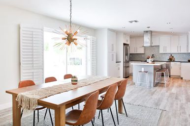 Sunlit open-concept dining and kitchen with a wooden table and tan leather chairs, starburst chandelier, kitchen island, stainless appliances, white cabinets and light wood floors.