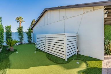 Sunny residential backyard with a mini artificial putting green, small golf flags, white slatted storage enclosure against the house, palm tree and tall hedges