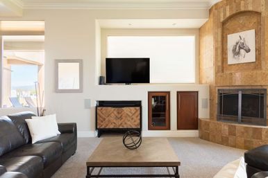 Bright contemporary living room with dark leather sofa, wooden coffee table topped with a metal orb, wall TV over a chevron wood cabinet, built-in cabinets and stone-tiled fireplace with horse artwork, and a sunny window overlooking a patio.