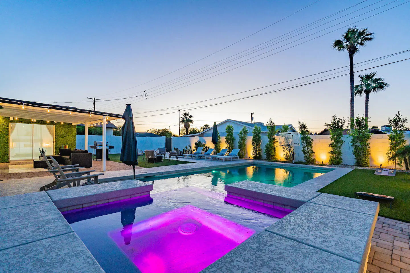 Dusk modern backyard with illuminated swimming pool and purple-lit spa, pergola with string lights, lounge chairs, and tall palm trees