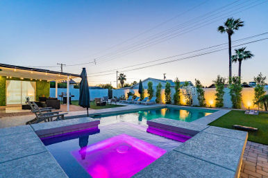 Dusk modern backyard with illuminated swimming pool and purple-lit spa, pergola with string lights, lounge chairs, and tall palm trees