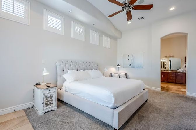 Bright modern master bedroom with tufted king bed and white bedding, high shuttered windows, ceiling fan, vintage bedside table, and open archway to an en-suite bathroom with double vanity