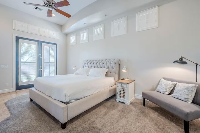 Bright contemporary master bedroom with tufted upholstered bed, neutral bedding, carpeted floor, bedside tables and lamps, gray loveseat, ceiling fan, clerestory windows and French doors with shutters letting in natural light.