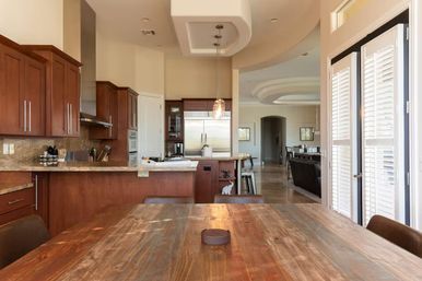 Sunlit open-plan kitchen and dining area with a rustic wooden table in the foreground, cherry wood cabinets, granite countertops, stainless refrigerator and hanging pendant lights.