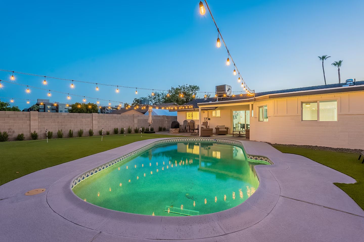 Glowing residential backyard pool at dusk with hanging string lights, patio lounge seating, and palm trees
