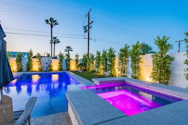 Resort-style backyard at dusk with a glowing rectangular pool and purple-lit spa, palm trees, lit privacy hedges, small lawn, poolside chairs and a poolside basketball hoop.