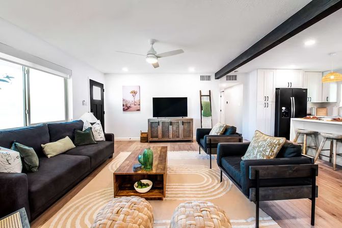 Bright open-concept living room and kitchen with dark sofa and matching armchairs, wooden coffee table, woven poufs, patterned area rug, TV on a wood console, and a breakfast bar with stools — modern suburban home interior.