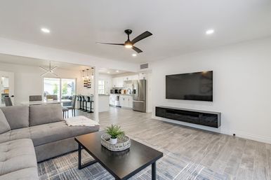 Bright modern open-concept living room and kitchen with gray sectional sofa, black coffee table, wall-mounted TV, ceiling fan, stainless-steel fridge and breakfast bar with stools near sliding glass door.
