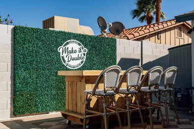 Sunny southwestern backyard patio with a wooden outdoor bar, four woven bistro bar stools, an artificial boxwood hedge wall with a round 'Make it a Double' sign, cinderblock fence and palm tree
