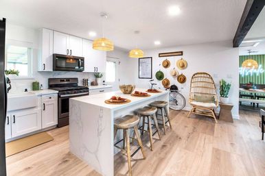 Bright coastal-inspired open kitchen with marble waterfall island, three rustic wood stools, rattan pendant lights, white cabinets, hat wall and wicker chair