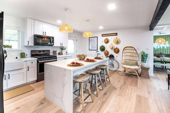 Bright coastal-inspired open kitchen with marble waterfall island, three rustic wood stools, rattan pendant lights, white cabinets, hat wall and wicker chair
