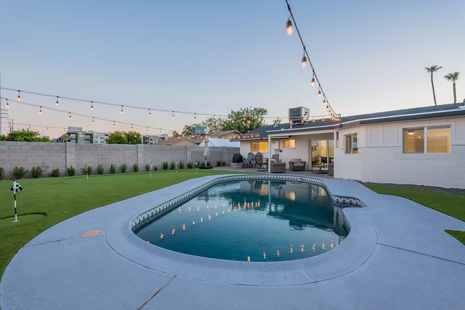 Suburban backyard at dusk with a kidney-shaped in-ground pool, string bistro lights, outdoor lounge seating and a small putting green framed by a block wall and palm trees