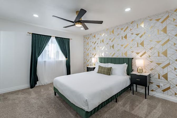 Stylish modern bedroom interior with emerald green upholstered headboard and curtains, crisp white bedding, gold geometric accent wallpaper, black ceiling fan, and matching bedside tables with lamps.