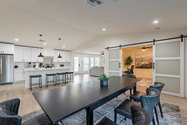 Modern open-concept kitchen and dining area in a bright home — white cabinets, stainless steel fridge, island with stools, black pendant lights, long black dining table with quilted chairs, and sliding barn doors to a cozy office.