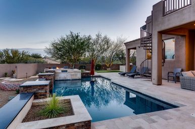 Modern Southwestern backyard at dusk with rectangular swimming pool and raised spa, stone tile patio, spiral metal staircase to an upper deck, lounge chairs, outdoor fire feature and desert landscaping