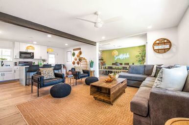 Bright open-concept living room and kitchen with hardwood floors, gray sectional sofa, leather armchairs, wooden coffee table, woven rattan light fixtures, and a green dining nook with neon wall sign.