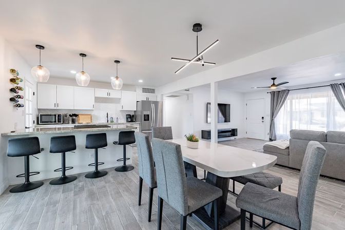 Bright modern open-concept kitchen and living room with white cabinets, stainless steel appliances, marble-look countertop, breakfast bar with four black stools under three glass pendant lights, adjacent dining table with gray upholstered chairs, light wood-look tile floor and a gray sofa facing a wall-mounted TV.