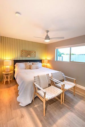 Bright modern boho bedroom with white quilted bed, sunburst wall art, rattan bedside lamps, two woven leather-strap chairs at the foot, ceiling fan and large window over wood-look flooring