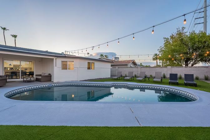 Cozy suburban backyard with a kidney-shaped pool at dusk, string lights overhead, patio seating near a single-story home, lounge chairs on green turf and palm trees in the background.