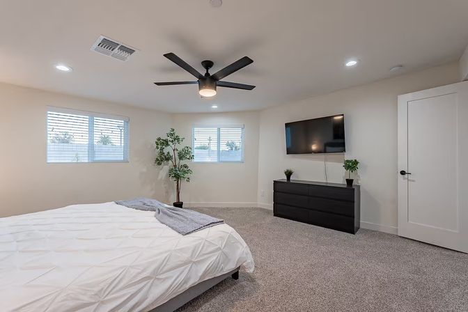 Spacious modern bedroom with white walls and neutral decor, king bed with white duvet and gray throw, large black ceiling fan, wall-mounted flat-screen TV above a black dresser, two windows with blinds, potted plants, and plush gray carpet.