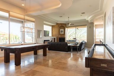 Sunlit modern game room and living area in a luxury home with a pool table, shuffleboard, leather sofas, fireplace and large windows overlooking a landscaped yard.