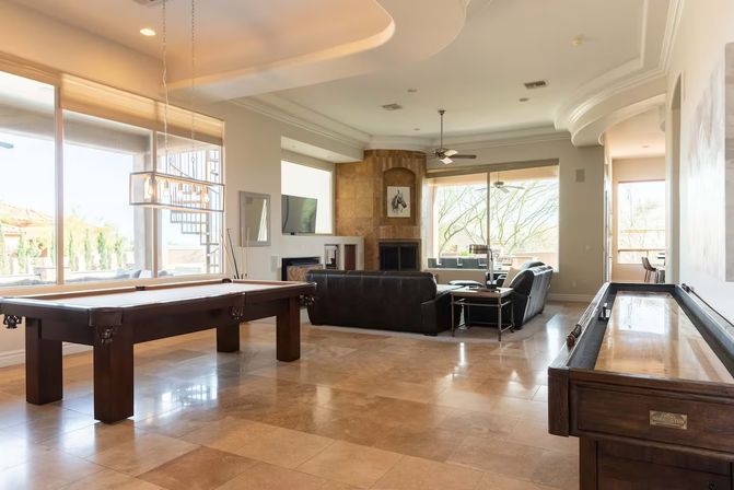 Sunlit modern game room and living area in a luxury home with a pool table, shuffleboard, leather sofas, fireplace and large windows overlooking a landscaped yard.
