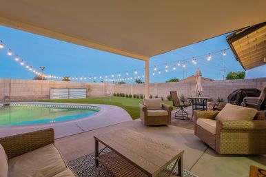 Covered suburban backyard patio at dusk with wicker lounge chairs and coffee table, curved swimming pool, artificial turf lawn, patio dining set and warm string lights overhead.